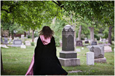 It's midday and Linda, clothed in a black cape with a pink hood, is walking away from us, into cemetary