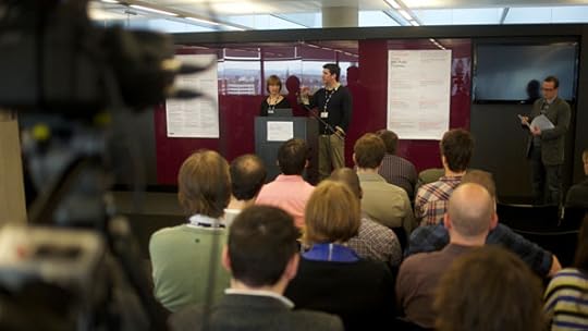 Two people speaking in a large open-plan meeting room. A video camera is out of focus in the foreground.