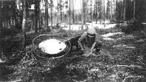 A trapper scrapes a fresh skin, as a beaver hide stretches on a frame nearby.