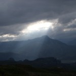 picture of the sun shining through a break in the clouds with a mountain in the background