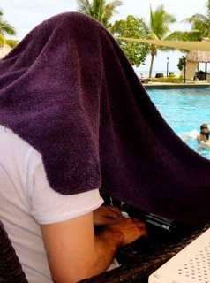 Man with head under towel typing on computer, while at beach resort.