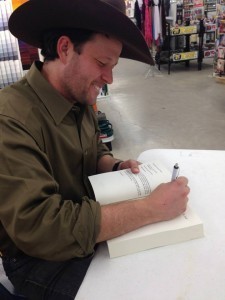 Philip Chavanne, smiling an wearing a cowboy hat, sings a copy of his novel in a book store