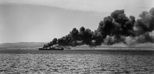 Operation Husky, invasion of Sicily, 10 July 1943: four miles south of Syracuse in 'Acid North' sector, an ammunition supply ship hit by enemy bombs burns close to the assault beaches. Landing craft can be seen on the shoreline below the smoke. Empire Elaine  offloaded her landing craft at Acid North that morning and may well be one of the ships lying beyond the burning vessel in this photograph (Lt F.G. Roper, R.N., Admiralty Official Collection, IWM A 18091).