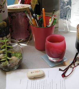 desk with cup of pencils, printed manuscript, pink glass candle, pair of glasses, eraser, award, small terrarium