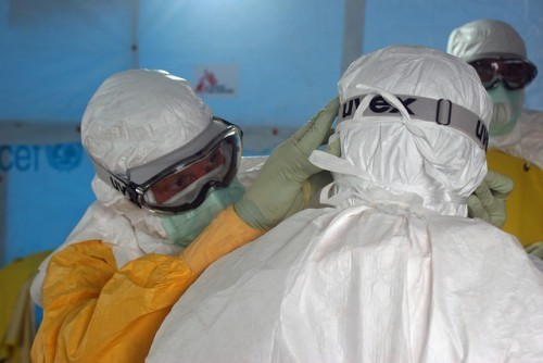 Dr. Joel Montgomery, Team Lead for CDC’s Ebola Response Team in Liberia, adjusts a colleague’s [personal protective equipment] before entering the Ebola treatment unit. Photograph by Athalia Christie. Public domain via Flickr.