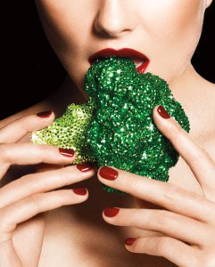 A beautiful shirtless woman bites into raw broccoli that's encrusted with glitter and rhinestones. 