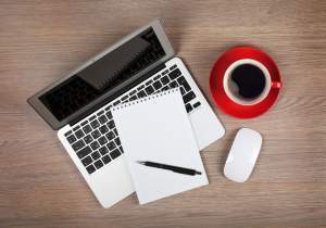 Blank notepad over laptop and coffee cup on office wooden table