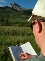Man reading a book outside with a mountain view in the distance.