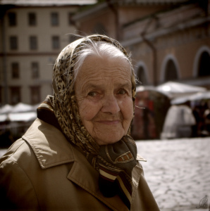 An old woman with a head scarf, smiling slightly. A wisp of hair escapes her scarf. 