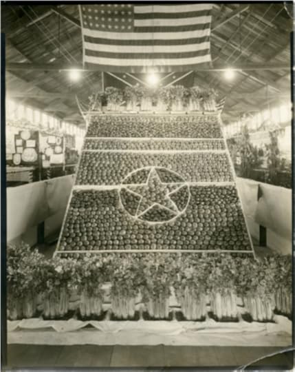 Photographic print. A large produce display, at the New York State Fair was created using a frame with horizontal spaces and a star within a circle in the center. The pattern within the frame was created with tomatoes and sweet peppers. In front of and on the top of the slanted display are bunches of celery. An American flag hangs above the display. Surrounding the display are decorated booths featuring more produce and educational presentations. 