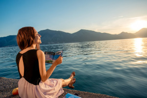 Young woman reading book and looking at beautiful sunrise on the pier with sea and mountains on background