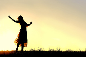 A woman wearing a long skirt, with long blonde hair, is dancing and praising God, while silhouetted against the evening sky