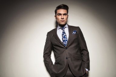 Young elegant business man leaning on a grey wall with his hands in pockets, looking away from the camera.
