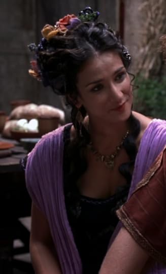 Actress Indira Varma, in purple robes, with her hair half up and little flowers and fruits and things stuck in it. 