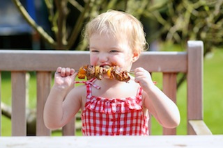 Funny toddler girl eating tasty grilled meat outdoors