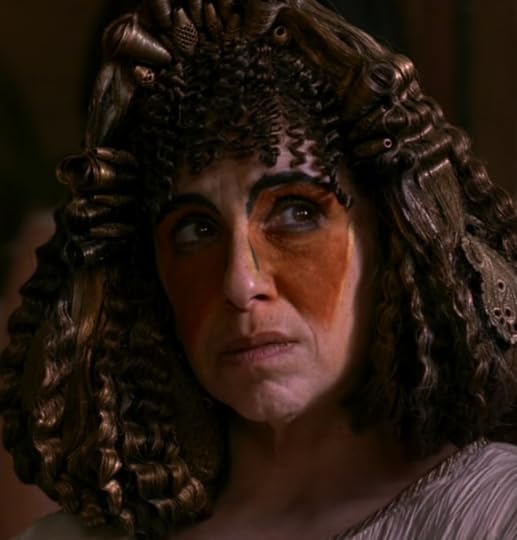 A middle-aged lady wearing a really busy wig with beads and starchy curls, with very exaggerated black eyebrows drawn on and triangles of rust-colored makeup over her eyes.