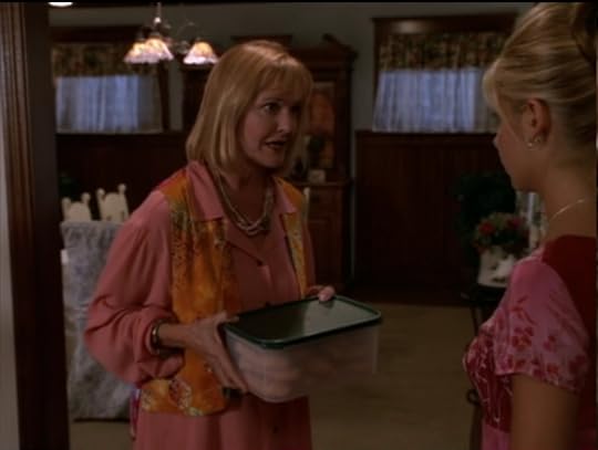 This woman has a shoulder-length blonde bob with bangs, a pink blouse with puffy sleeves, an orange batik silk vest on over that, a necklace that's like, multiple ropes of either small shell or wood beads, and a copper bracelet. 