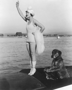 1916: American actor Gloria Swanson (1899 - 1983) stands on tiptoes on the prow of a motorboat while Teddy the dog sits with his paws on the steering wheel in a still from director Clarence G Badger's film 'Teddy at the Throttle'.