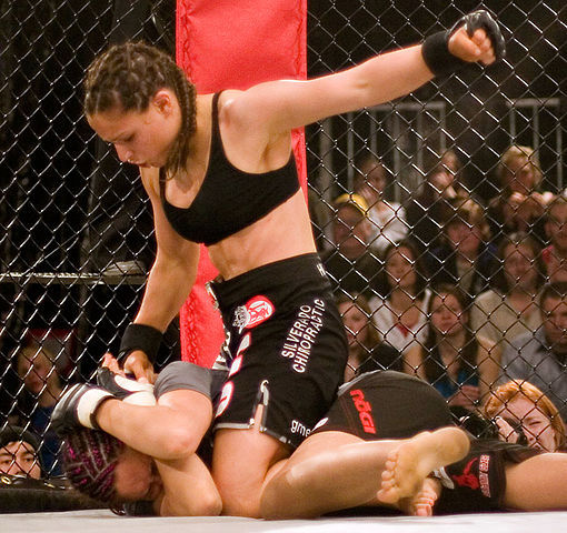 Gina Carano in full mount position performing ground and pound. Photo by Matthew Walsh. CC BY-SA 2.0 via Wikimedia Commons.