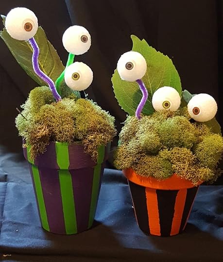 Two terracotta flower pots. One of them is painted with black and orange stripes. The other is painted with purple and green. Both of them contain mounds of moss and a fake leaf. Styrofoam balls with googly eyes on them are stuck on pipe cleaners that are stuck in the moss, giving the appearance of eyes growing on stalks like plants.