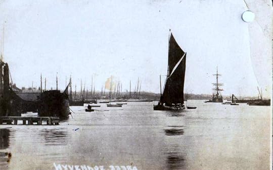 A black and white photograph of the River Colne at Wivenhoe. A Thames barge in full sail is in the middle of the photograph; a square rigger is to the right in the distance. On the left, the masts of many boats can be seen, and a jetty is in the foreground.