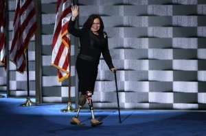 Sen. Lamma Tammy Duckworth, a half-Asian-American woman, walks onto a stage waving with one hand, holding a cane in the other, in front of an American flag. She wears a black suit. She has two prosthetic legs, one with an American flag on it.