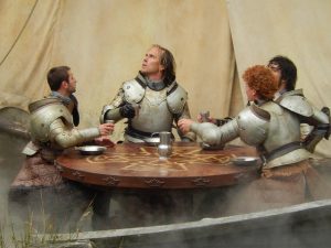 Four knights in armor (without their helmets) sit around a table with metal plates set before them. They are all white men, differentiated primarily by the color of their hair.
