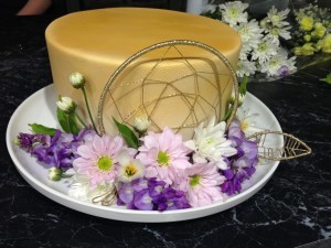 Front shot of cake, a single-tier 23cm round cake covered in fondant (with sharp corners!), sprayed gold, with a glittery gold dream catcher centered on the front side of the cake (it extends above the edge of the cake by about a centimetre); three sprays of purpley-blue wisteria are arranged around the bottom of the cake at the front on the white platter; they are filled out with three baby pink daisies, three white daisies, some white daisy buds, a few wisteria leaves, and a gold glittery feather or leaf made to match the dreamcatcher.