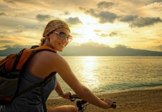 Young woman with backpack standing on the shore near his bike and smiling.
