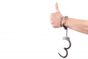 a white man's hand, in one of a pair of handcuffs, gives the viewer a thumbs-up
