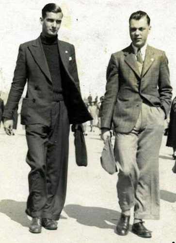 Man on left wearing dark turtle neck with dark jacket and pants; man on right wearing lighter jacket and pants.