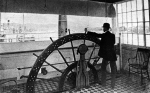 A pilot stands at the wheel in the pilothouse of an unknown riverboat