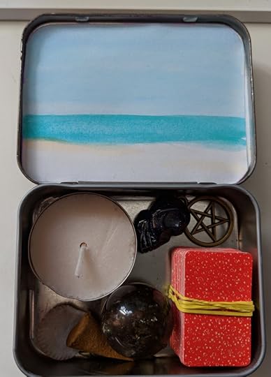 A photo of my purse-sized altar, a converted Sucrets tin with a tealight candle, a smoky quartz sphere, a small stone skull, a brass pentacle, a very, very tiny deck of tarot cards, a seashell, and a cone of incense. The inside of the lid of the tin has a water color scene of the ocean