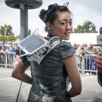 Kitty Yeung stands with her back to the camera looking over one shoulder, displaying her 3D printed solar panel wearable recharging station on her back.