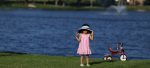 young girl wearing a pink dress and large hat standing near a tricycle in front of a pond with fountain