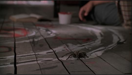 A warped, unfinished wooden floor and a portion of the painted symbol. A tarantula is moseying his way along the perimeter.
