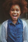 African-American female child with her mouth open in a wide smile wearing a blue denim button up shirt with white crochet sweater over top