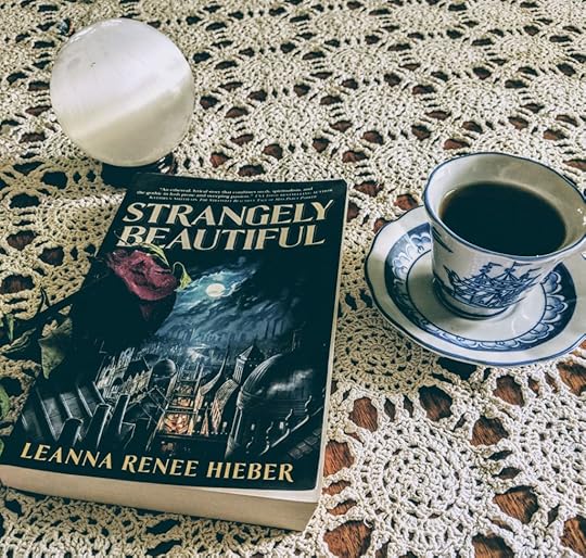 A photo of the book Strangely Beautiful by Leanna Renee Hieber, staged on my crocheted white table cloth with a china cup of tea with saucer beside it, a dried rose resting across it, and a selenite crystal ball looking ghostly and beautiful above it.