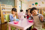 children sitting at a table with bowls of chalk in front of them wearing smocks in a school setting