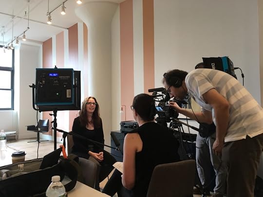 Laurie, sitting in a chair and facing a recording team. Their backs are to us. On the left is a large light, then a young woman with dark hair seated talking to Laurie, then a man wearing headphones bent over a camera screen.