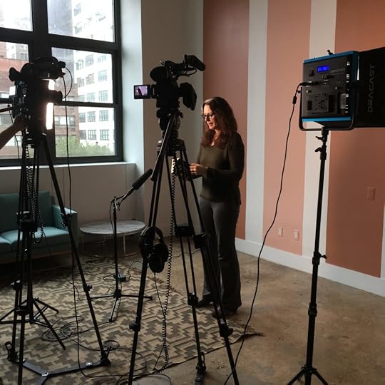 Laurie stands against a wall painted in stripes of dark beige and white, wearing a green sweater and gray pants. Several cameras are set up around her, and she's about to read from SHOUT.