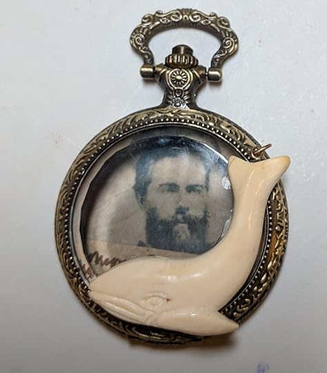A pocket watch with a glass lid. Inside is a photo of Herman Melville. An ivory whale charm is affixed to the front edge of the glass.