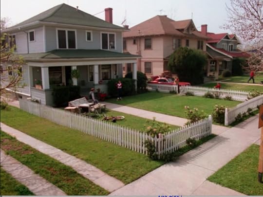 A gray house at the end of a row of houses, all two-story with small front lawns and white picket fences. The gray house has what appears to be a full living room set on the damn lawn.