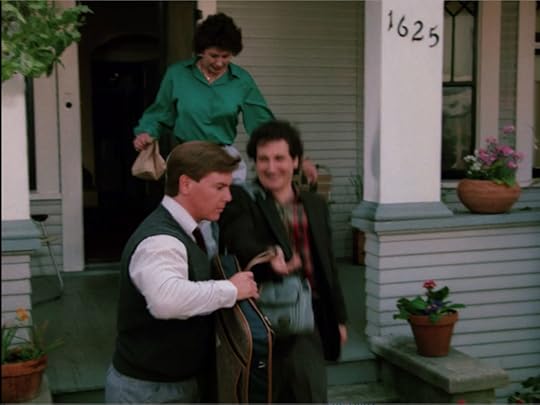 A slightly motion-blurred shot of Linn-Baker's character, Larry, heading down the porch steps of the gray hous. He's got a camera bag over his shoulder and is followed by a woman who is clearly meant to be his mother, as she carries a sack lunch. Another adult man, this one in a sweater vest and button down like he's wandered in from a movie about young Wall Street hustlers hands Larry a garment bag.