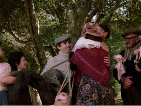 Balki is hugged by a woman in a kerchief, red shawl, and floral print skirt. A girl in a peasant blouse stands to the left of the shot, with another woman. A young man in a turtle-neck sweater and jaunty cap are behind them. An old man with glasses, a newsboy cap, and a scarf stands to the right