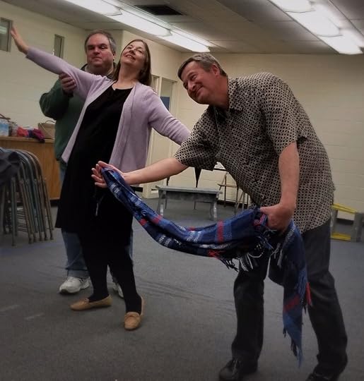 Photo of three people rehearsing a play. A man stands behind a woman, she has her arms spread wide and she is smiling. Another man is dancing with a blanket as if it is his partner.