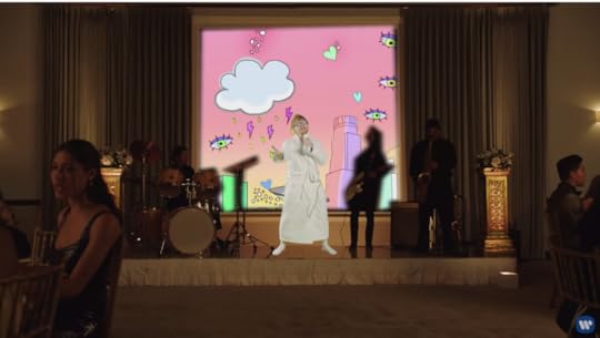 Ed is on a stage singing in a bathrobe and white socks, in front of a backdrop of a pink sky with falling clouds, hearts, and eyes.