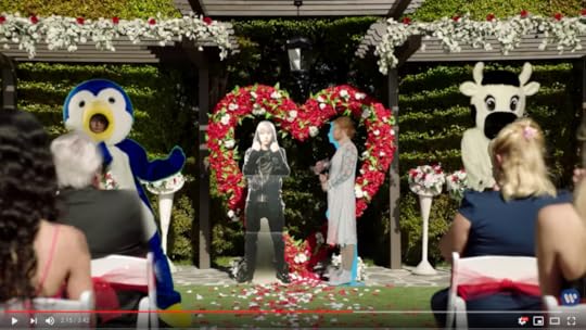 Where to begin. Okay, the overall scene is a wedding. Ed Sheeran in a penguin suit dances on one side of the screen, while the cow mascot dances on the right. In the middle, Ed Sheeran in a wedding dress stands under a heart-shaped arch of roses, marrying a Justin Bieber cut-out.