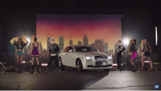 Ed and Justin are both dressed by cowboys. They flank a white Bentley. There are sexy video girls dancing on either side of them, and there is a painted backdrop of a city skyline at sunset.