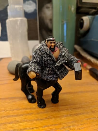 A small centaur toy with a black horse body, tan man body, goatee, and little painted-on crown. Bronwyn Green sewed him a tiny houndstooth business jacket and given him a briefcase.